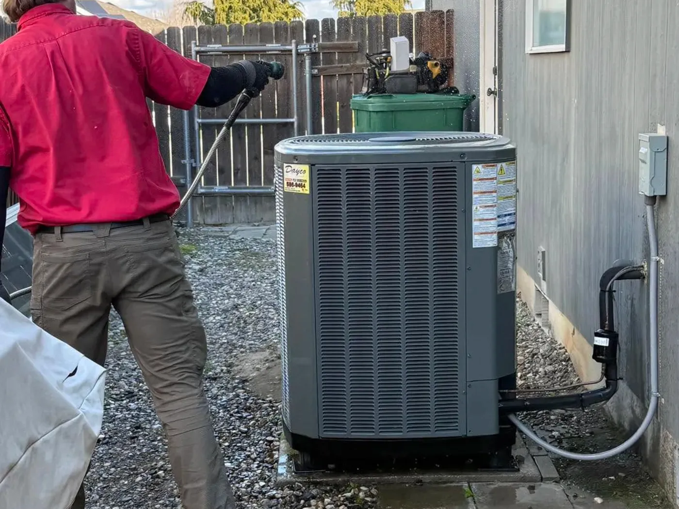 A Dayco technician in a red company shirt standing beside an outdoor air conditioner