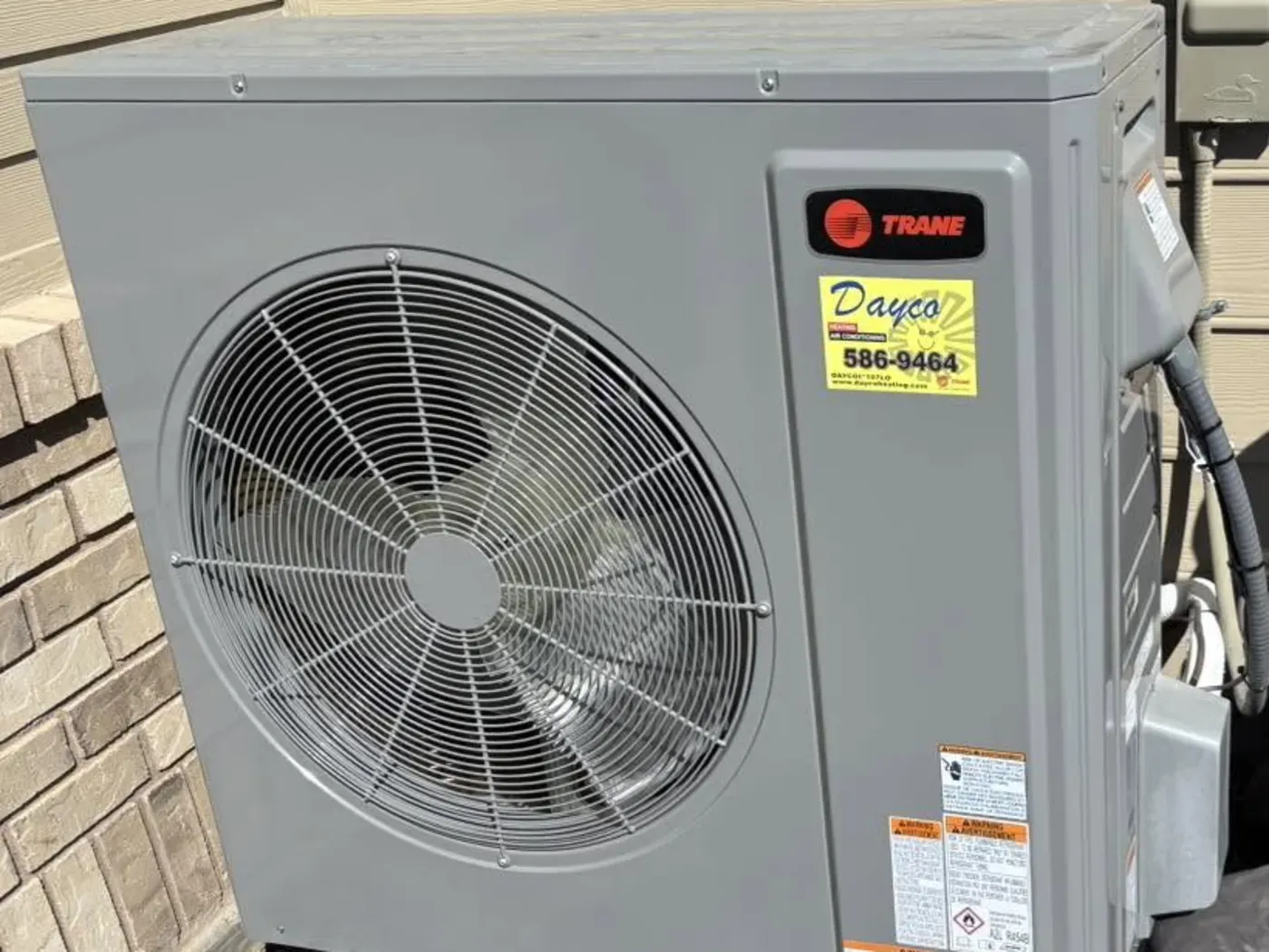 A new Trane outdoor unit with a Dayco service sticker, installed against a brick house