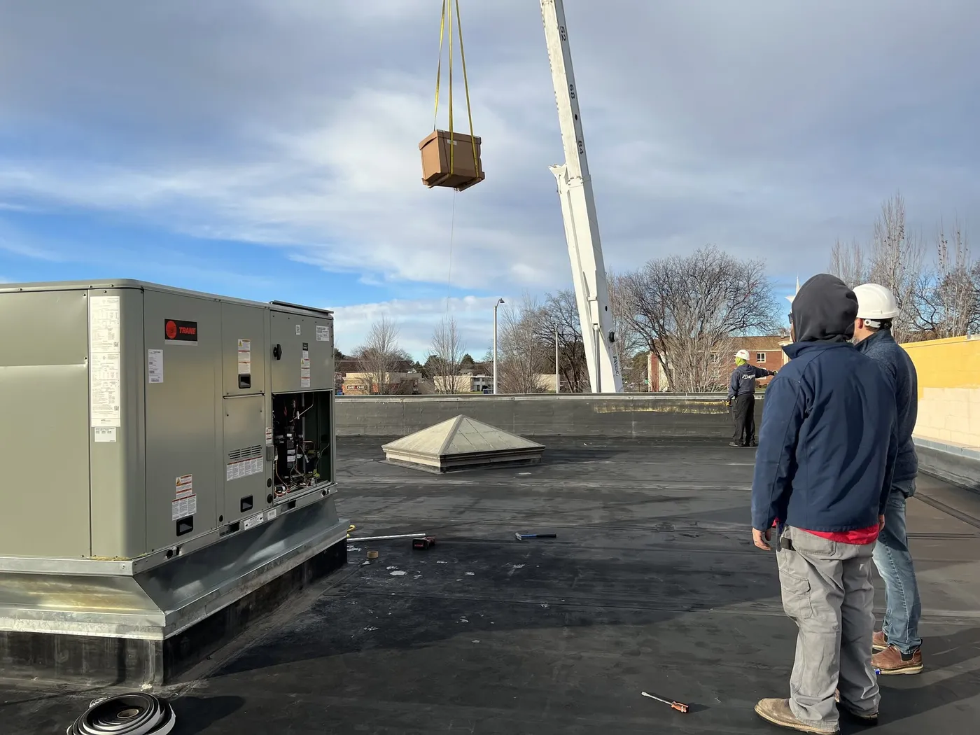 A crane lowers a new packaged rooftop unit toward a commercial roof while two Dayco crew members in hard hats watch beside an installed Trane unit