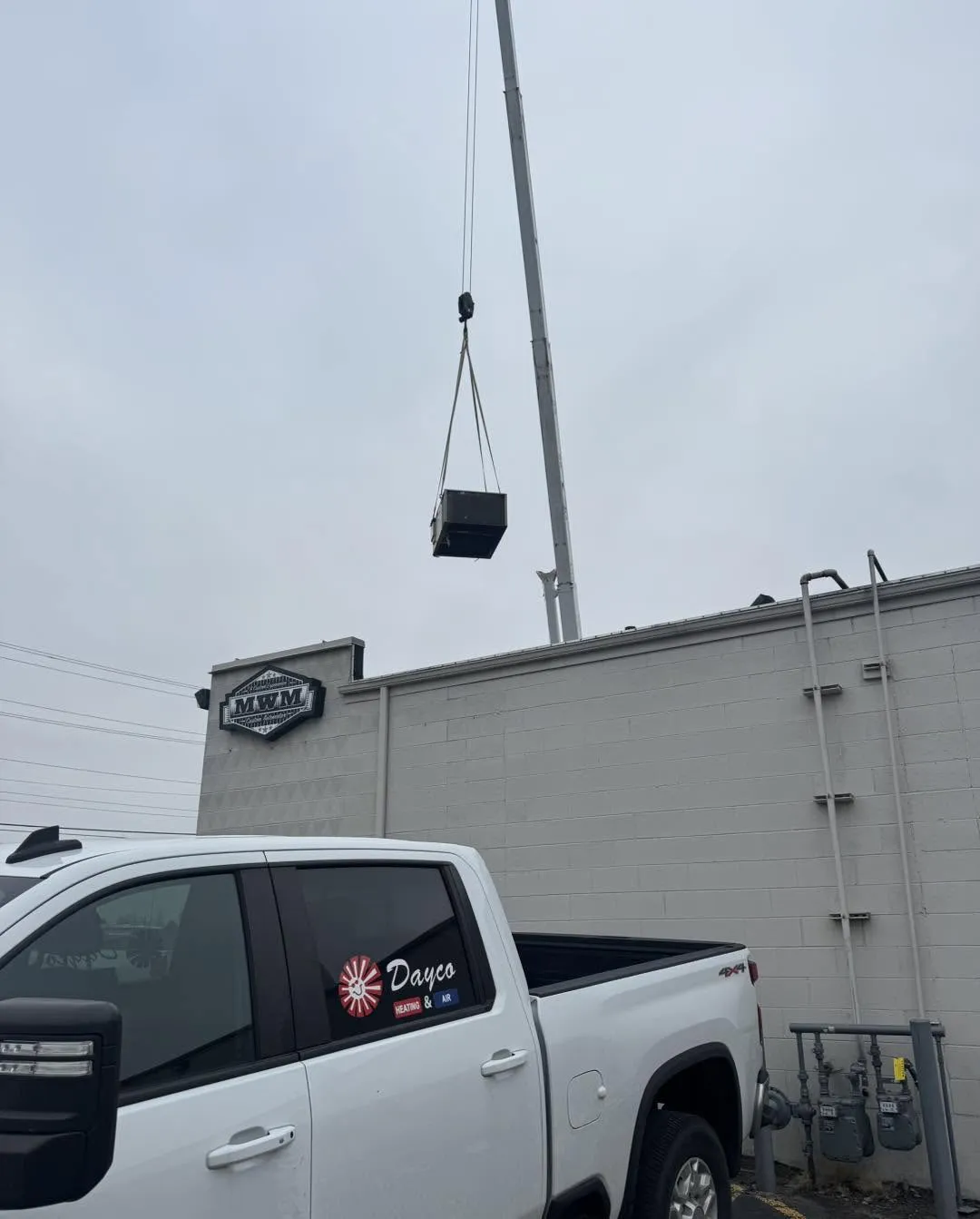 A crane lifts a new rooftop unit onto a commercial building while a Dayco pickup waits below