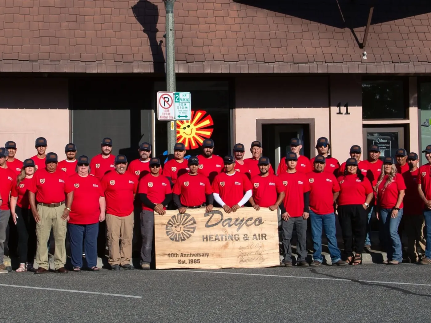 The Dayco crew in red company shirts lined up outside the shop at 11 N Auburn in Kennewick, holding a carved wooden sign that reads “Dayco Heating & Air, 40th Anniversary, Est. 1985”